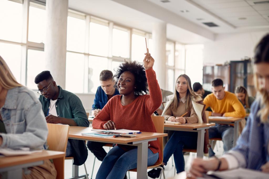 学生 of a variety of races sit at tables in a white pillared classroom with a row of windows against wall. An African American female student, 穿着长袖, 赭石的t恤, smiles and raises her hand.